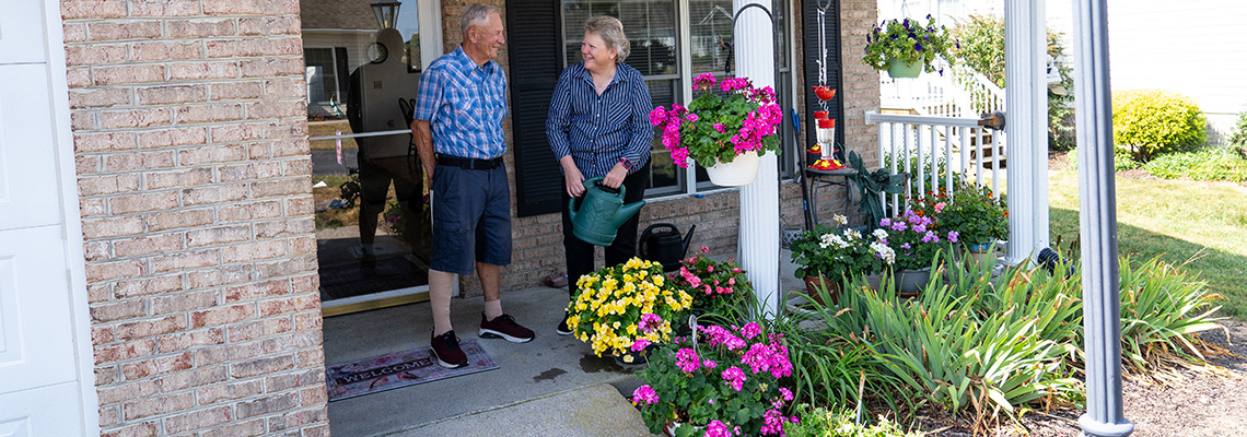Two people stand and talk on a porch surrounded by colorful flowers and plants; one holds a green watering can.