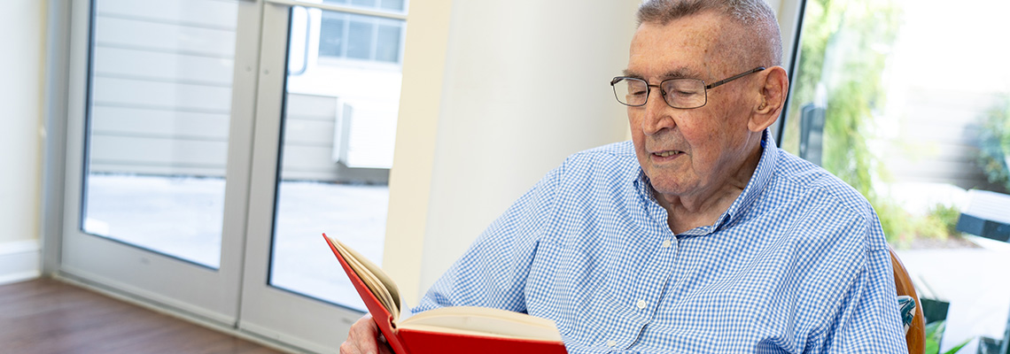 An elderly man wearing glasses and a blue checkered shirt sits indoors by a window, reading a red book.
