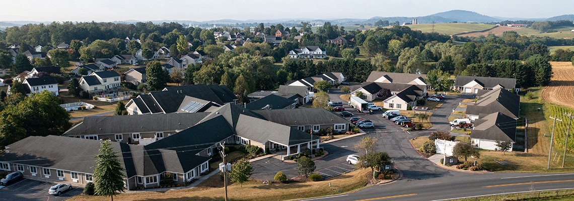 Aerial view of a suburban neighborhood with houses, a large building complex, parking lots, trees, and distant hills under a clear sky.